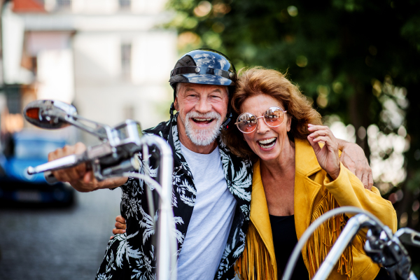 A front view of cheerful senior couple travellers with motorbike in town.