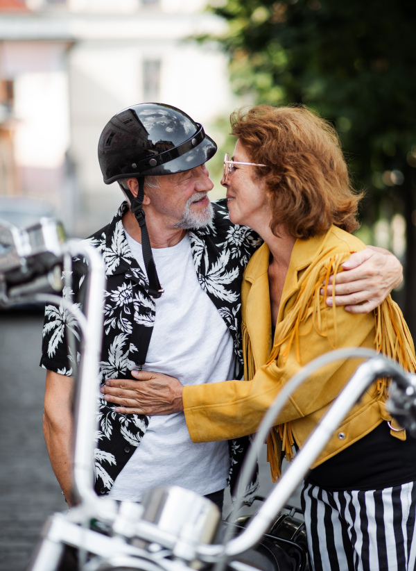 A front view of cheerful senior couple travellers with motorbike in town, kissing.