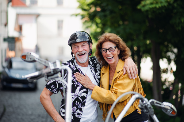 A front view of cheerful senior couple travellers with motorbike in town.