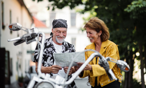 A front view portrait of cheerful senior couple travellers with motorbike in town, looking at map.