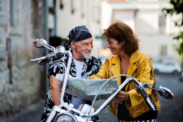 A front view portrait of cheerful senior couple travellers with motorbike in town, looking at map.