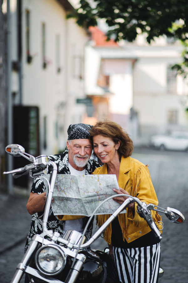 A front view portrait of cheerful senior couple travellers with motorbike in town, using map.