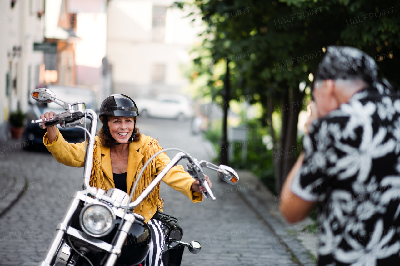 A front view of cheerful senior couple travellers with motorbike in town, taking photograph.