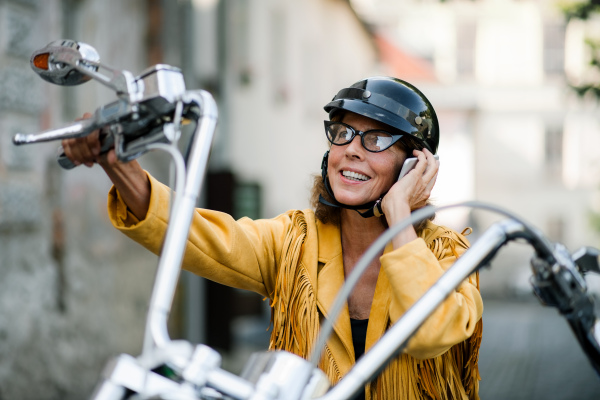 A cheerful senior woman traveller with smartphone and motorbike in town, making phone call.