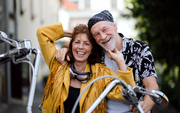 A front view of cheerful senior couple travellers with motorbike in town.