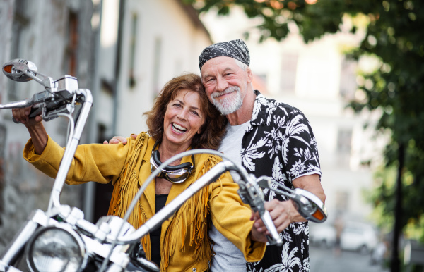 A front view of cheerful senior couple travellers with motorbike in town.