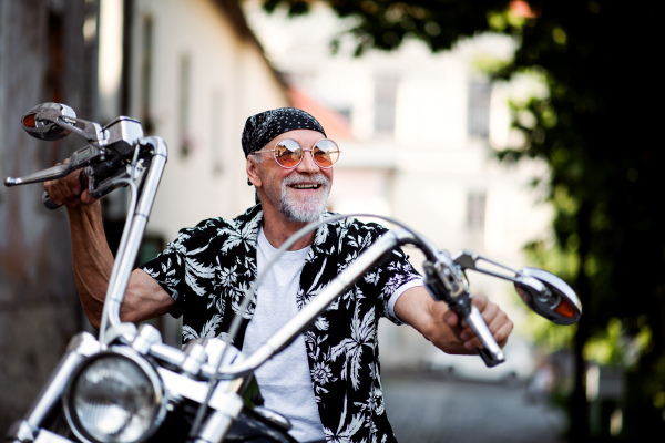 A front view of cheerful senior man traveller sitting on motorbike in town.