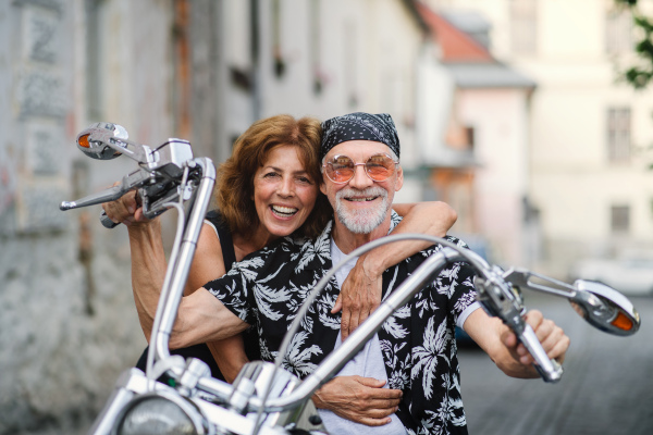 A front view of cheerful senior couple travellers with motorbike in town.