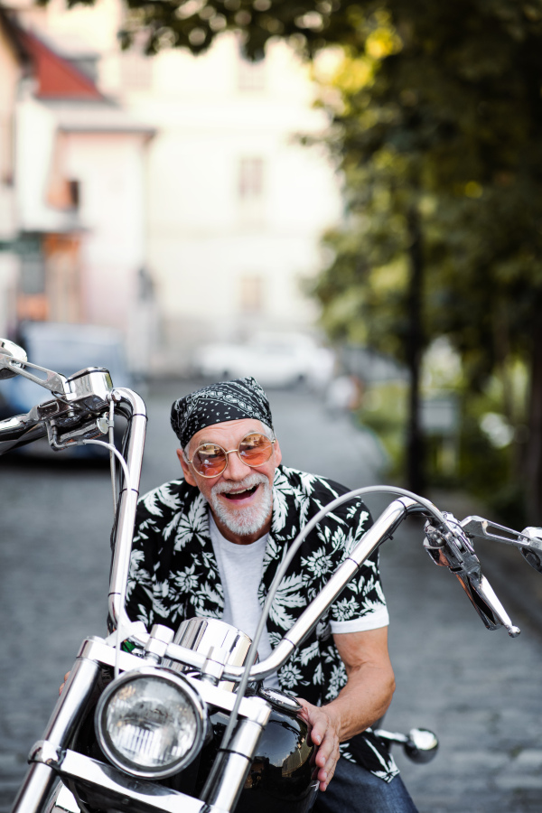 A front view of cheerful senior man traveller sitting on motorbike in town.