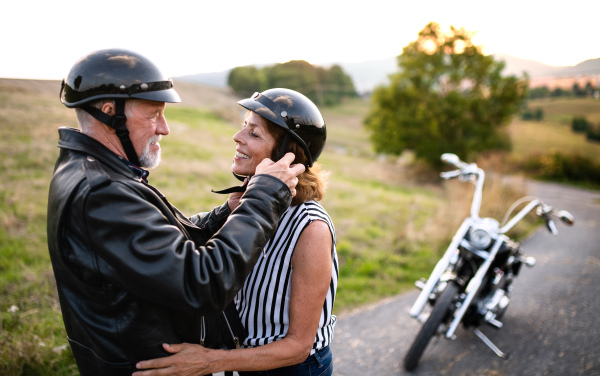 A front view portrait of cheerful senior couple travellers in love with motorbike in countryside.