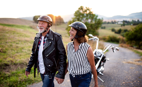 A front view portrait of cheerful senior couple travellers with motorbike in countryside.