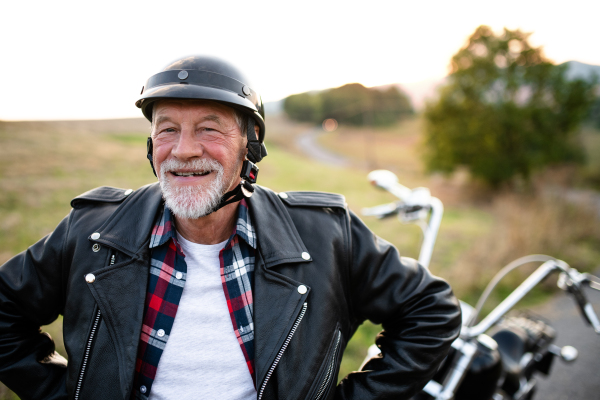 A front, view portrait of cheerful senior man traveller with motorbike in countryside, standing.