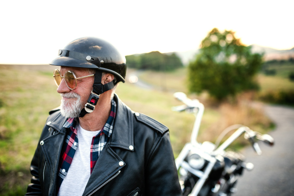 A front, view portrait of cheerful senior man traveller with motorbike in countryside, walking.