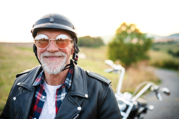 A front view portrait of cheerful senior man traveller with motorbike in countryside, headshot.