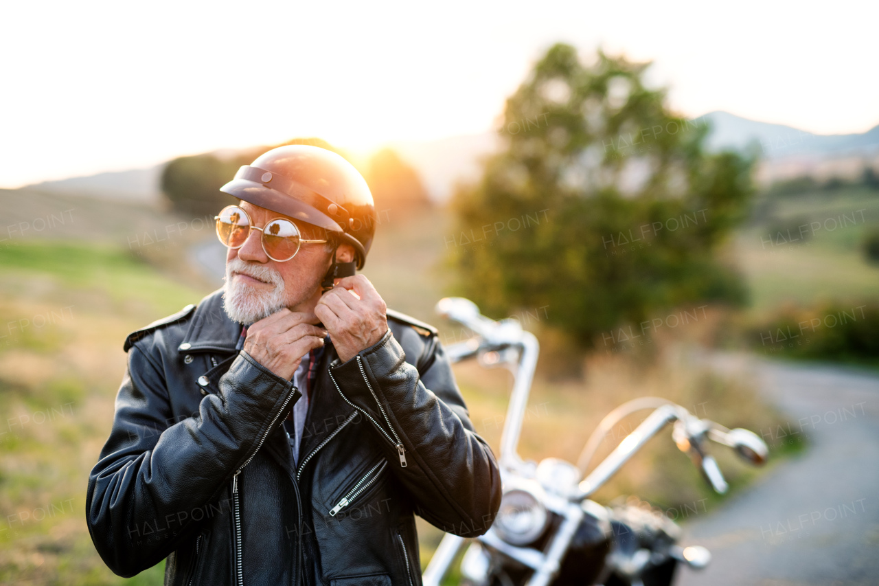 A senior man traveller with motorbike in countryside at sunset, putting on helmet.