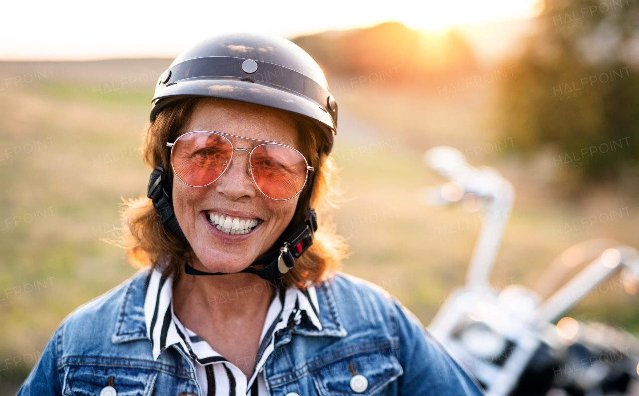 A cheerful senior woman traveller with motorbike in countryside at sunset, standing.