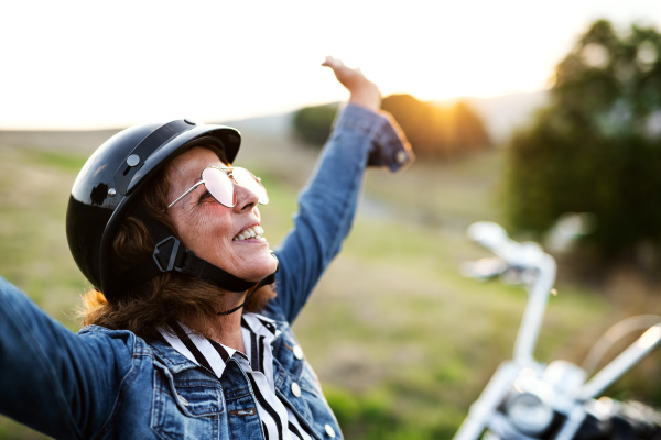 A cheerful senior woman traveller with motorbike in countryside, standing.