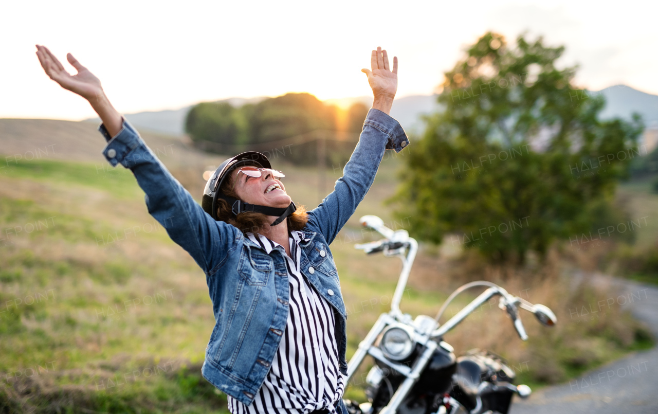 A cheerful senior woman traveller with motorbike in countryside, standing.