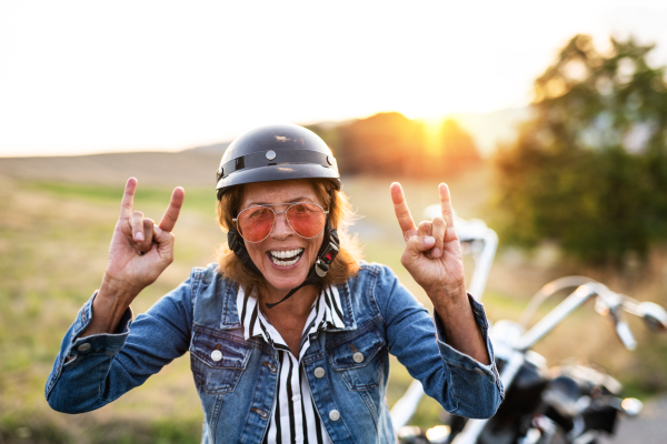 A cheerful senior woman traveller with motorbike in countryside at sunset, standing.