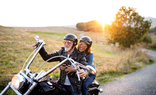 A cheerful senior couple travellers with motorbike in countryside at sunset.