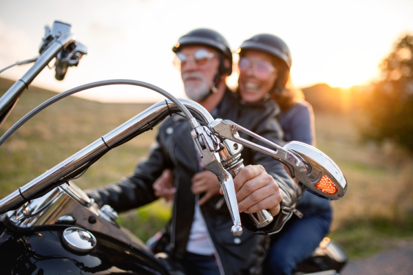 A cheerful senior couple travellers with motorbike in countryside at sunset.