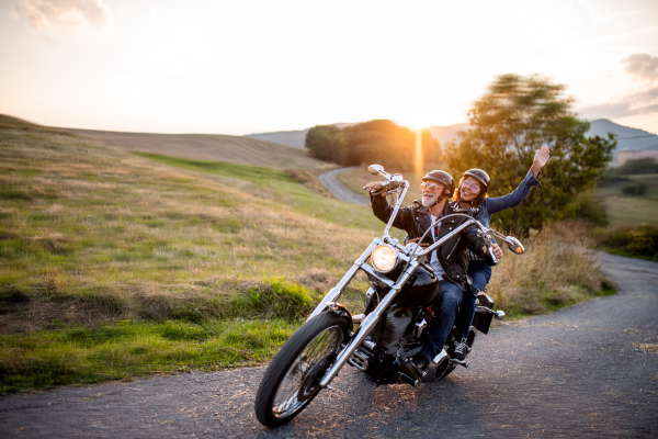 A cheerful senior couple travellers with motorbike in countryside at sunset.