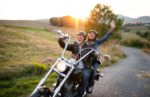 A cheerful senior couple travellers with motorbike in countryside at sunset.