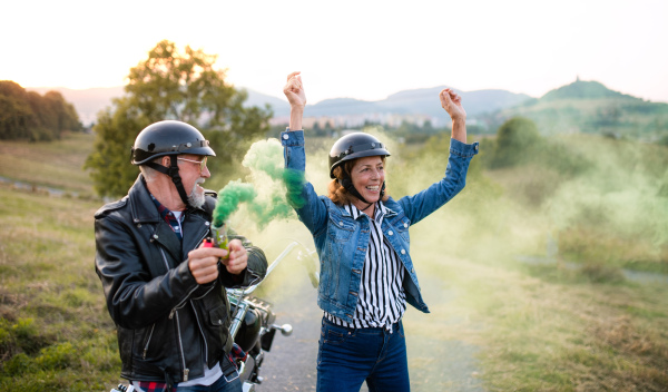 A front view portrait of cheerful senior couple travellers with motorbike in countryside, having fun.