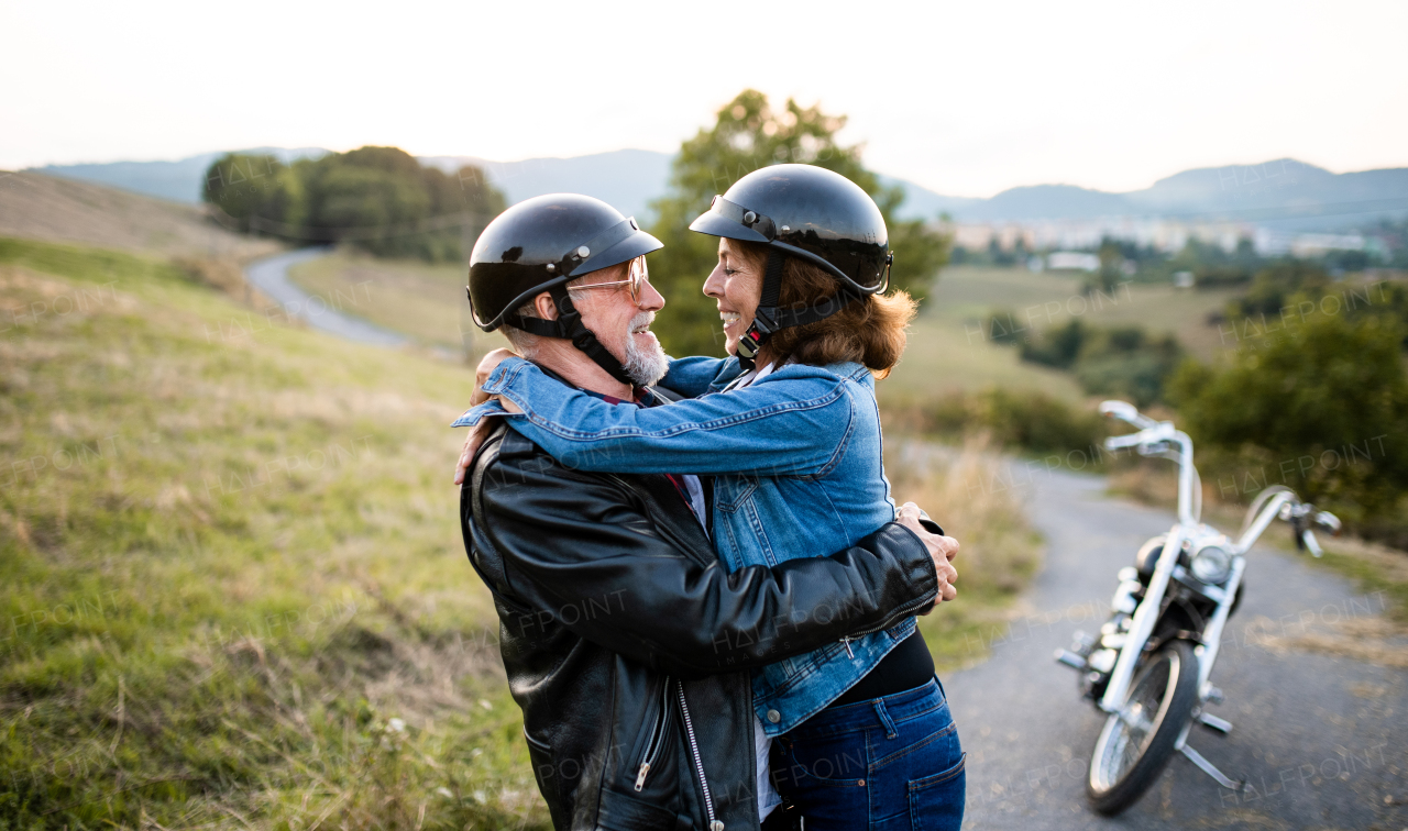 A front view portrait of cheerful senior couple travellers with motorbike in countryside, hugging.