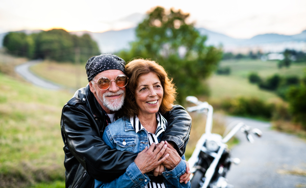 A front view portrait of cheerful senior couple travellers with motorbike in countryside.