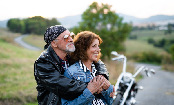 A front view portrait of cheerful senior couple travellers with motorbike in countryside.