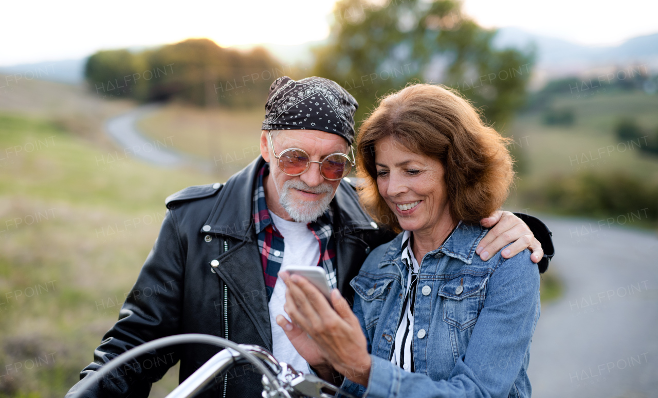 A cheerful senior couple travellers with motorbike in countryside, using smartphone.