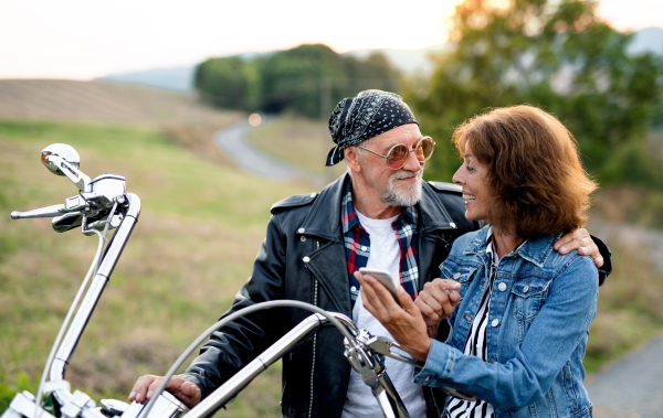 A cheerful senior couple travellers with motorbike in countryside, using smartphone.