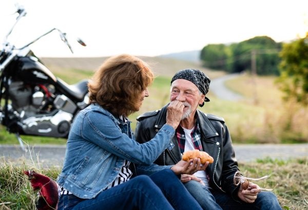 A portrait of cheerful senior couple travellers with motorbike in countryside, eating snack.