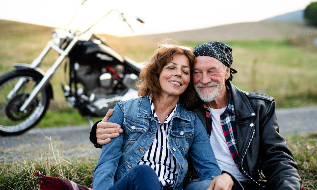 A front view of senior couple travellers with motorbike in countryside, resting.