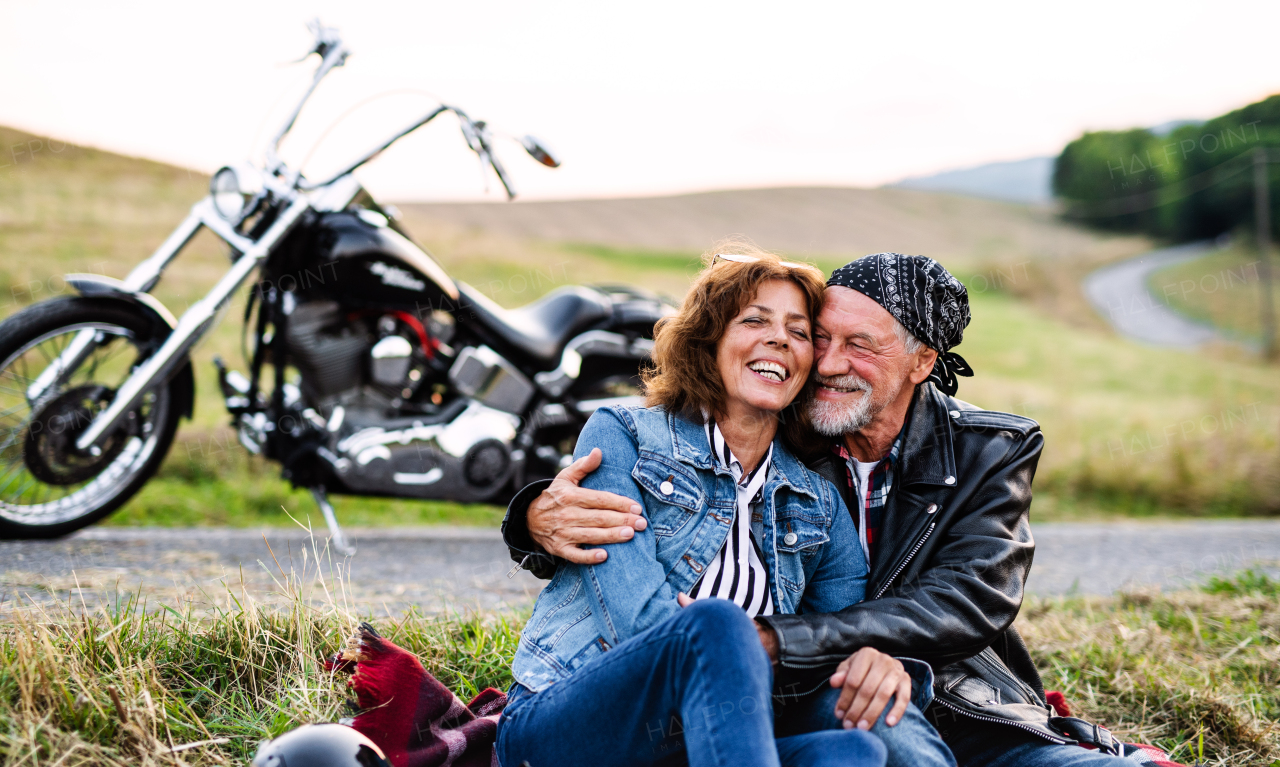 A front view portrait of cheerful senior couple travellers with motorbike in countryside.