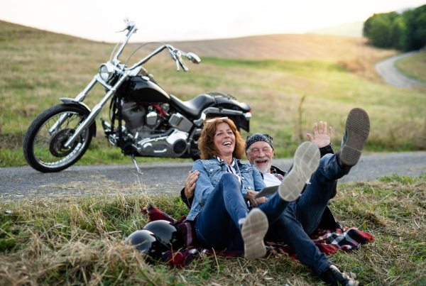 A front view portrait of cheerful senior couple travellers with motorbike in countryside.