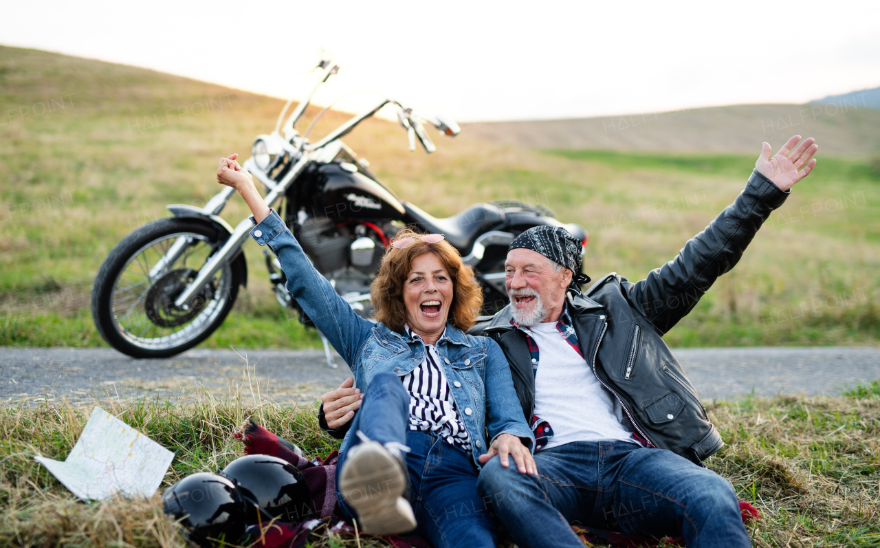 A front view portrait of cheerful senior couple travellers with motorbike in countryside, having fun.