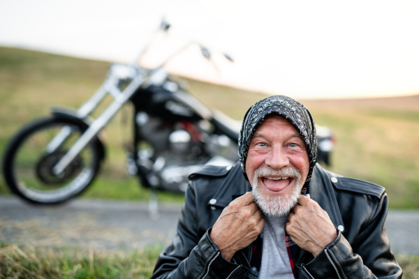 A front view portrait of cheerful senior man traveller with motorbike in countryside, having fun.