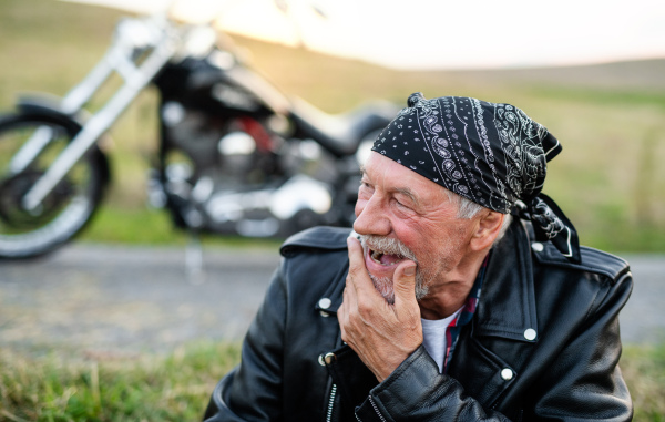 A front view portrait of cheerful senior man traveller with motorbike in countryside, resting.