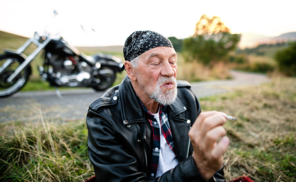 A portrait of cheerful senior man traveller with motorbike in countryside, smoking.