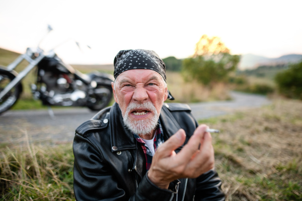 Angry senior man traveller with motorbike in countryside, smoking cigarette.