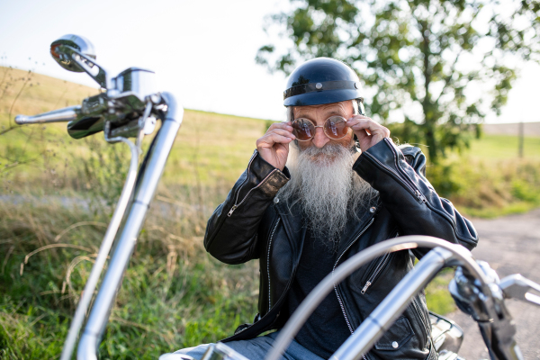 A senior man traveller with motorbike in countryside, putting on sunglasses.