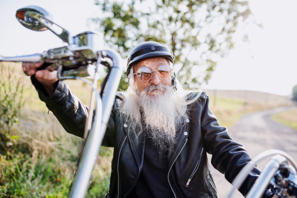 A senior man traveller with motorbike and sunglasses in countryside, looking at camera.