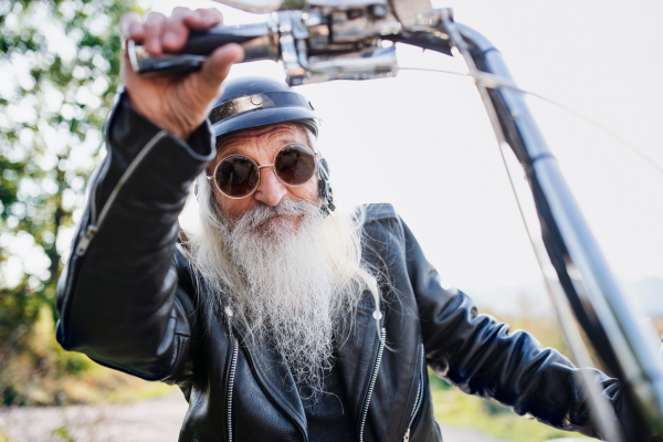 A senior man traveller with motorbike and sunglasses in countryside, looking at camera.