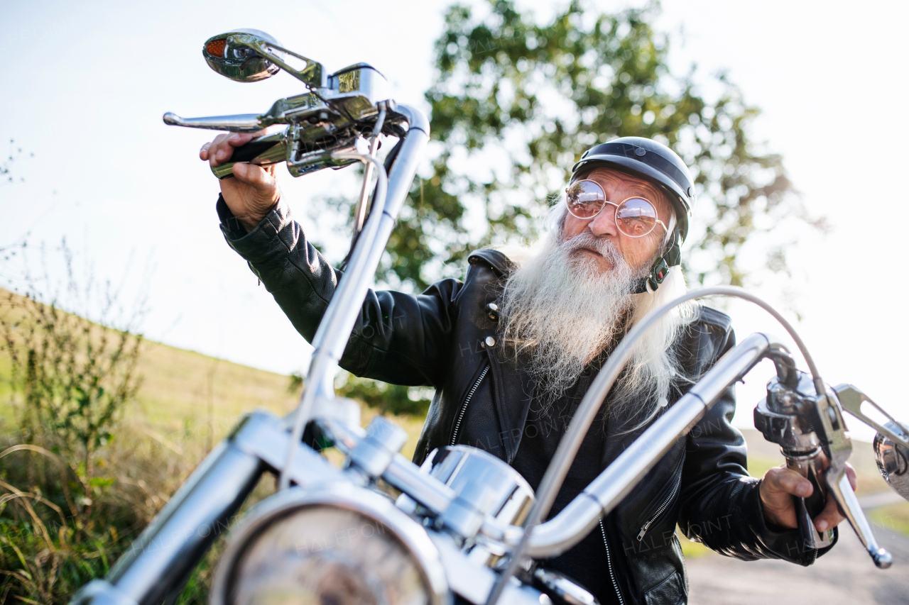 A senior man traveller with motorbike and sunglasses in countryside, looking at camera.