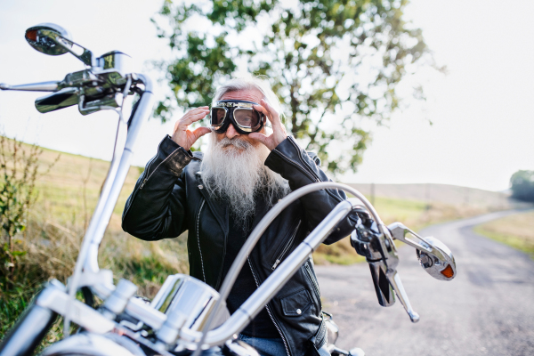 A senior man traveller with motorbike and goggles on road in countryside. Copy space.