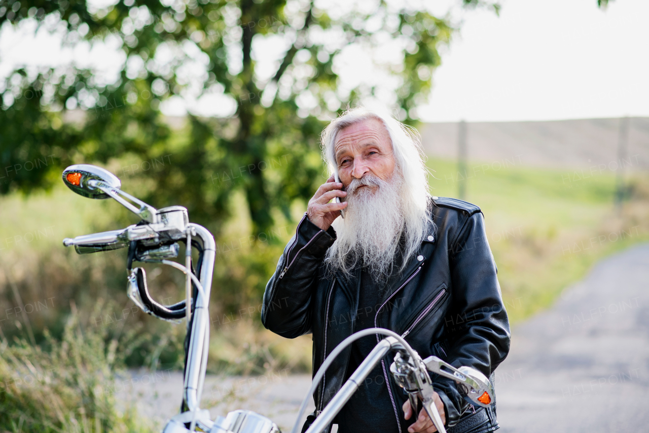 A senior man traveller with motorbike in countryside, using smartphone.