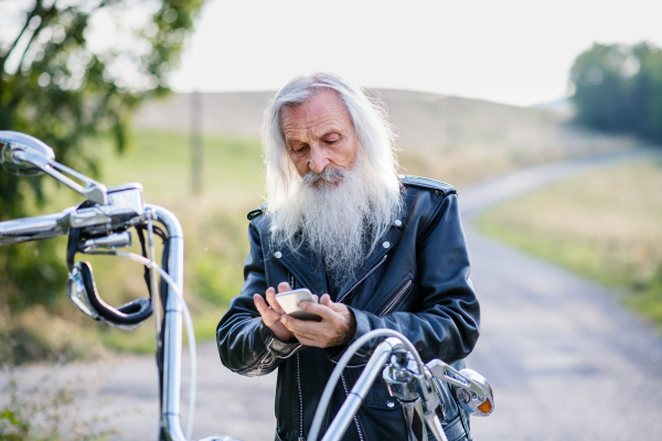 A senior man traveller with motorbike in countryside, using smartphone.