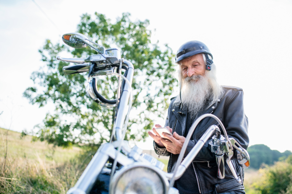 A senior man traveller with motorbike in countryside, using smartphone.